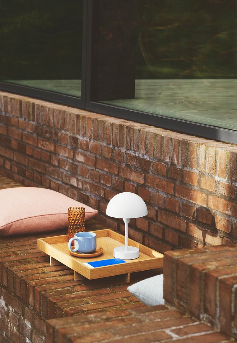 Holztablett mit einem blauen Akzent, einer weißen Lampe, einer blauen Tasse auf einem runden Untersetzer und einem bernsteinfarbenen Glas, auf einer Ziegeloberfläche mit Kissen.