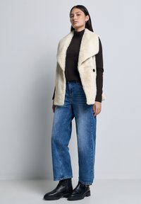 Gilet in finta pelliccia color crema con colletto a revers, abbinato a un dolcevita nero attillato e jeans in denim blu larghi, completato da stivaletti neri alla caviglia.