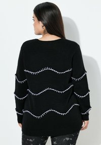 Schwarzer Strickpullover mit wellenförmigen weißen Nähten über den Rücken. Gerippter Kragen und Bündchen. Lockere Passform. Weiche Textur.