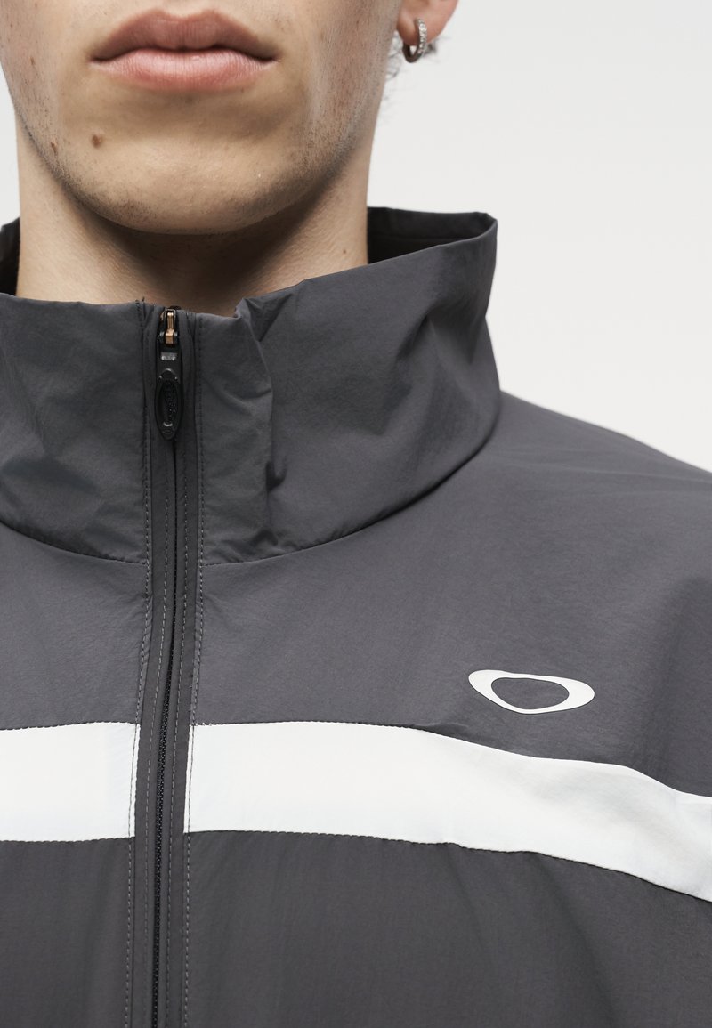 Primer plano de una persona que lleva una chaqueta gris oscuro con una franja horizontal blanca y un pequeño logo circular cerca del hombro.