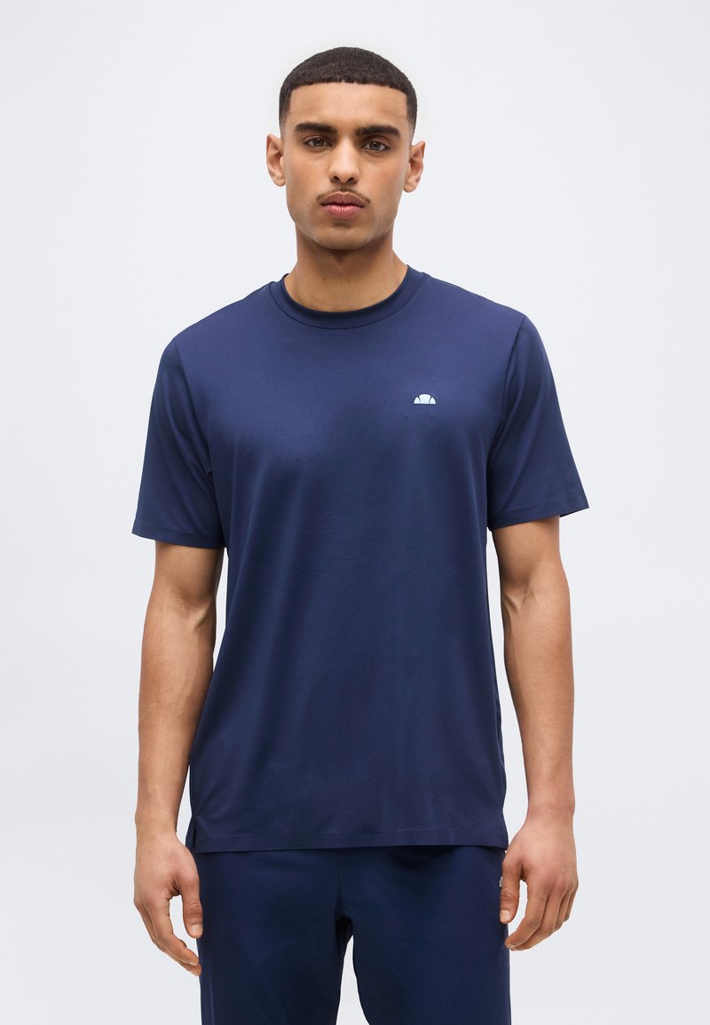 Νεαρός άνδρας με κοντά μαλλιά, ντυμένος με μπλε σκούρο πουκάμισο με κοντά μανίκια και παντελόνι στο ίδιο χρώμα, στέκεται μπροστά σε απλό φωτεινό φόντο.