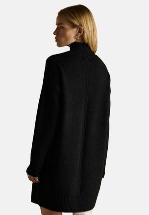 Frau mit blonden Haaren, die ein schwarzes, hochgeschnittenes, langärmliges Strickkleid trägt und nach links vor einem einfachen Hintergrund schaut.