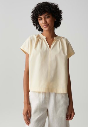 Femme aux cheveux bouclés portant un chemisier jaune pâle à manches courtes et un pantalon blanc, debout devant un fond gris uni.