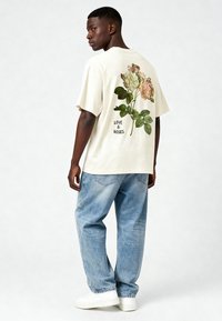 Bēša grafiskā t-kreklā ir ziedu dizains uz muguras ar uzrakstu "MĪLESTĪBA UN ROŽES". Apvienots ar zilu džinsu biksēm un baltām sporta apaviem.