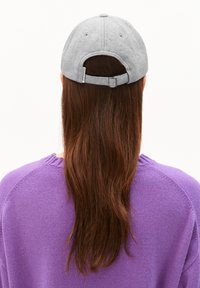 Des cheveux bruns longs portés lâchement sous une casquette de base-ball gris claire, associés à un pull en laine violet, montrés de dos sur un fond blanc.