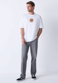 Hvid t-shirt med korte ærmer og et rundt orange og rødt grafik, parret med grå jeans og sorte sneakers med hvide detaljer.