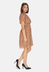 Vestido floral marrón con mangas cortas, cintura entallada y falda acampanada; lo lleva con tacones de aguja negros. Tela suave con una textura delicada.