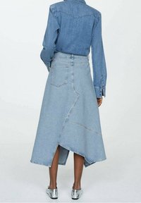 Gonna di jeans, blu chiaro, di lunghezza media con orlo asimmetrico, caratterizzata da cuciture evidenti e tasche. Abbinata a una camicia di jeans con bottoni.