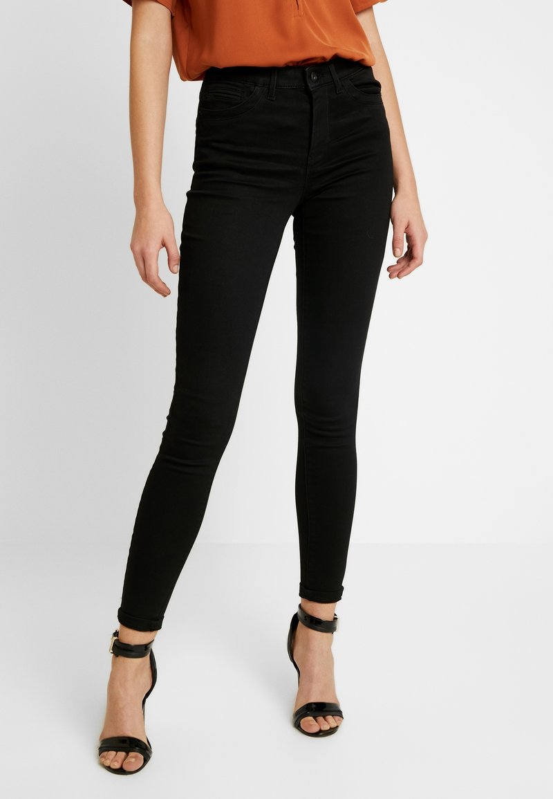 Vaqueros pitillo negros de cintura alta, hechos de tela de algodón elástica. Presentan cinco bolsillos y detalles de costura sutiles. Se llevan con tacones negros.