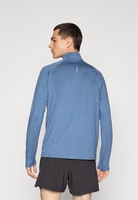 Cămașă sport albastră cu mâneci lungi, guler înalt, material texturat și detaliu reflectorizant pe spate, purtată cu pantaloni scurți negri.