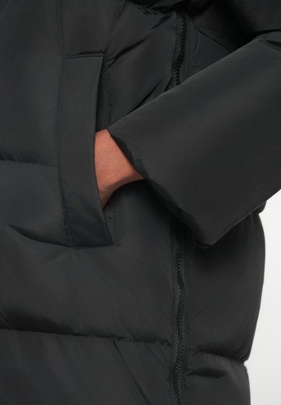Μαύρο μπουφάν με φερμουάρ μπροστά, υφασμάτινη υφή και πλαϊνή τσέπη με το χέρι να είναι partially ορατό μέσα.