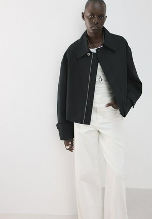 Personne portant une veste noire surdimensionnée par-dessus un haut blanc et un pantalon blanc à jambes larges, debout devant un fond blanc uni.