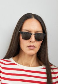 Occhiali da sole neri Ray-Ban con un design classico, caratterizzati da lenti tonde e un sottile ponte metallico. Indossati con una camicia a righe rossa e bianca.