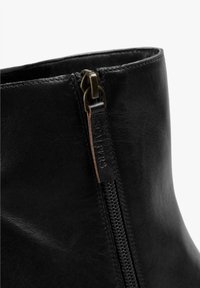 Botín de cuero negro hasta el tobillo con cremallera lateral, parte superior texturizada y una pequeña etiqueta de marca cerca de la cremallera como detalle.