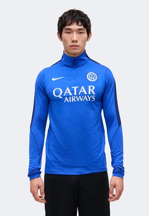 Mężczyzna w niebieskiej sportowej koszulce Nike z logo Inter Mediolan i sponsorem Qatar Airways, stojący na jednolitym jasnym tle.