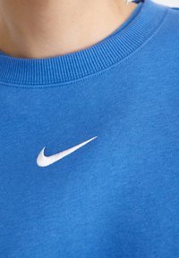 Närbild av en person som bär en blå t-shirt med rund hals och en liten vit Nike-swoosh-logotyp på bröstet.
