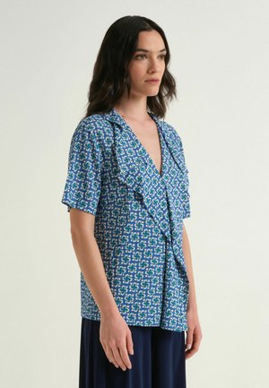 Femme portant un chemisier bleu à manches courtes à motif avec un col à volants, debout devant un fond clair uni.