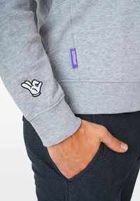 Sudadera gris con un acabado texturizado. Presenta un gesto de mano bordado en la manga y una etiqueta de marca morada en el lateral.