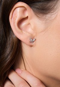 Srebrne kolczyki w kształcie motyla z przezroczystymi kamieniami, o prostych liniach i gładkich powierzchniach, zaprezentowane na ludzkim uchu.