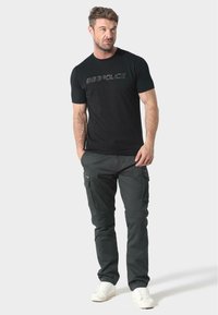 Černé tričko s vyraženým logem, spárované s tmavě šedými cargo kalhotami s kapsami a slim fit. Boty jsou bílé tenisky.