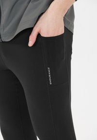 Schwarze sportliche Leggings aus dehnbarem Material, mit einer Seitentasche und dem "ENDURANCE"-Logo in Weiß auf dem Oberschenkel, in taillierter Passform.