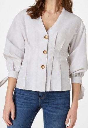 Kobieta ubrana w białą, zapinaną na guziki bluzkę z bufiastymi rękawami i związanymi mankietami, połączoną z niebieskimi dżinsami.