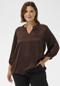 Smeđa satenska bluza s V-izrezom i naborima; ima rukave do tri četvrtine s elastičnim manžetama; opušteni kroj i mekani materijal.