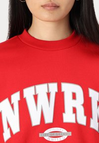 Felpa rossa con collo a coste, caratterizzata da audaci lettere bianche "NWRK" e un logo centrale con la scritta "Città originale dei sogni."
