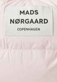 Doudoune rose rembourrée avec une étiquette en tissu blanc portant un texte noir indiquant "MADS NØRGAARD COPENHAGUE." Texture lisse, design minimal.