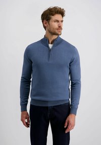 Blauer Strickpullover mit einem Reißverschlusskragen und gerippten Bündchen, kombiniert mit dunklen Jeans. Weiche Textur, taillierte Passform und klare Linien sind hervorstechende Merkmale.