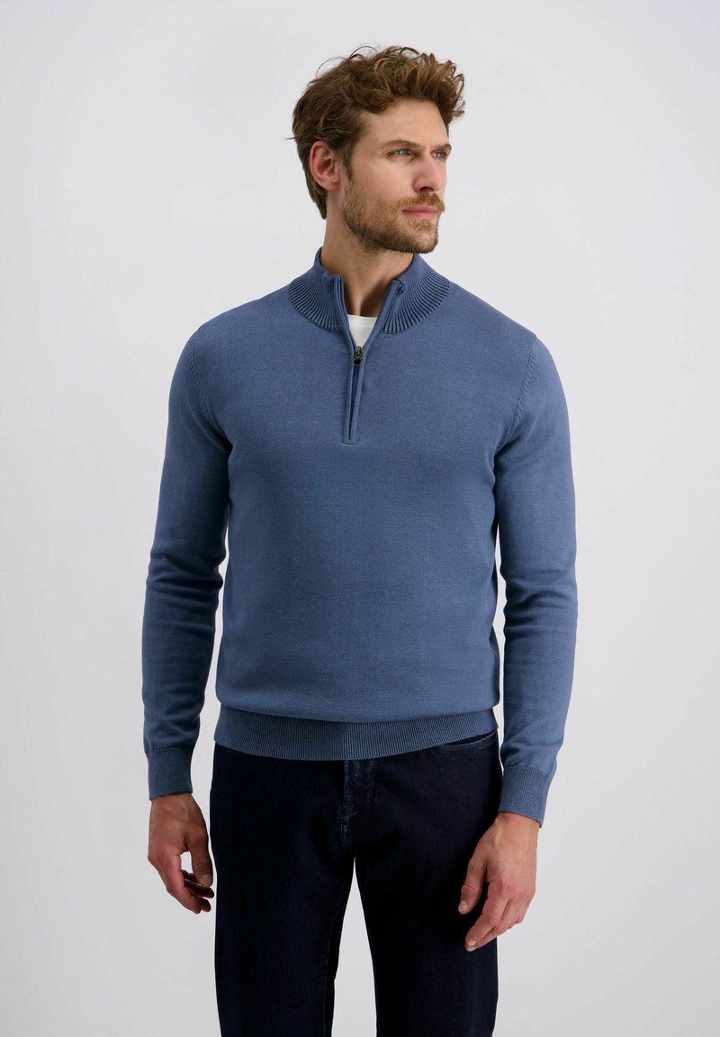 Blauer Strickpullover mit einem Reißverschlusskragen und gerippten Bündchen, kombiniert mit dunklen Jeans. Weiche Textur, taillierte Passform und klare Linien sind hervorstechende Merkmale.