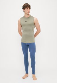 Αμάνικο αθλητικό τοπ σε ελαιόλαδο με μαύρο λογότυπο, συνδυασμένο με μπλε συμπιεστές κολάν. Λείος υφασμα και εφαρμοστή σχεδίαση.