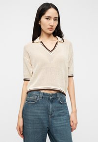 Pull polo en maille beige crème avec bordure marron, manches courtes et col en V. Associé à un jean en denim bleu taille haute.