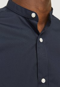 Navyblauw shirt met een opstaand kraag, voorzien van twee witte knopen bij de hals en een gladde, gestructureerde stof.