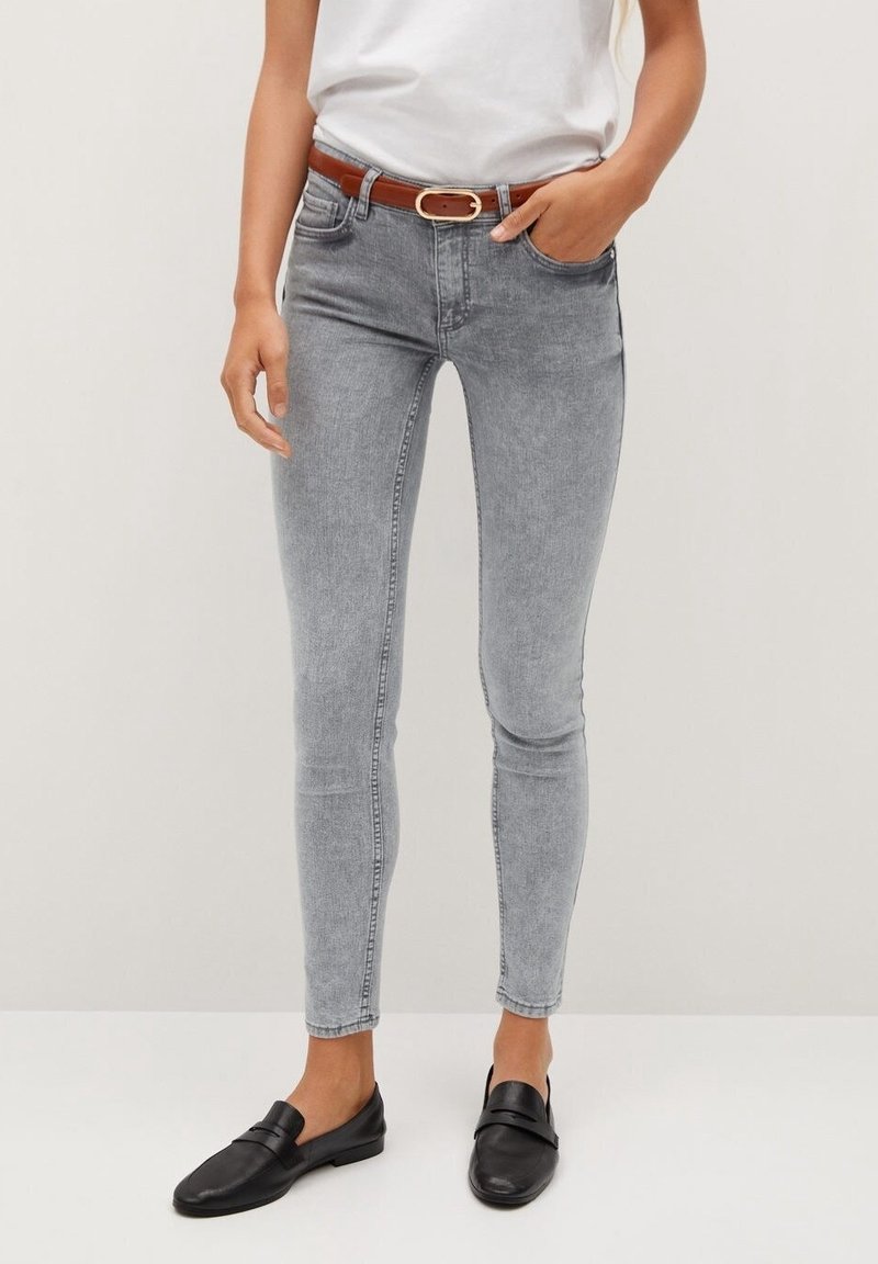 Femme portant un jean skinny gris clair avec une ceinture marron, un haut blanc et des mocassins noirs, debout devant un fond clair uni.