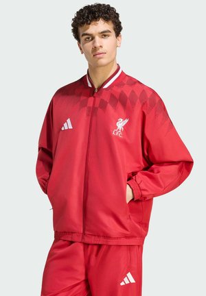 Junger Mann mit lockigem Haar, der einen roten Liverpool FC und Adidas Trainingsanzug trägt, steht mit den Händen in den Jackentaschen vor einfarbigem Hintergrund.