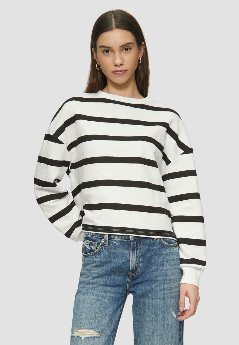 Felpa bianca con strisce orizzontali nere, scollo rotondo, vestibilità comoda e polsini a coste. Abbinata a jeans blu strappati.