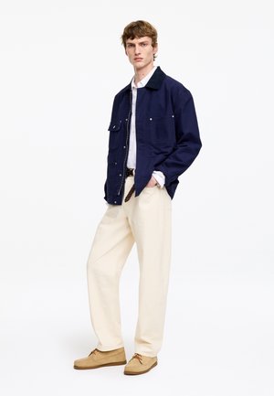 Junger Mann steht vor einem schlichten weißen Hintergrund und trägt eine marineblaue Jacke, ein weißes Hemd, cremefarbene Hosen und hellbraune Schnürschuhe.