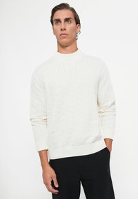 Maglione crema a maglia cable con colletto alto, caratterizzato da un motivo testurizzato, maniche a costine e orlo. Abbinato a pantaloni neri.