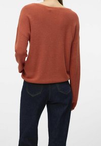 Femme portant un pull en maille couleur rouille et un jean bleu foncé, vue de dos contre un fond blanc uni.