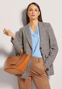 Braune Leder-Tragetasche mit flachen Griffen, glatter Textur und dezenten Stichdetails. Frau trägt ein kariertes Blazer über einer blauen Bluse.