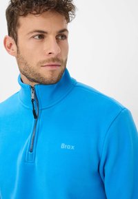 Pullover in pile blu con zip, caratterizzato da un alto colletto, un piccolo logo embossato sul petto e una zip in metallo con linguetta in tessuto.