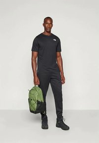 T-shirt preta leve, calças desportivas pretas e mochila verde com detalhes em rede; pessoa usa botas de caminhada pretas.