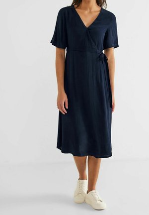 Dunkelblaues, knielanges Wickelkleid mit kurzen, flatternden Ärmeln, leichtem Stoff und Taillendetail mit Bindeband. Kombiniert mit cremefarbenen Turnschuhen.