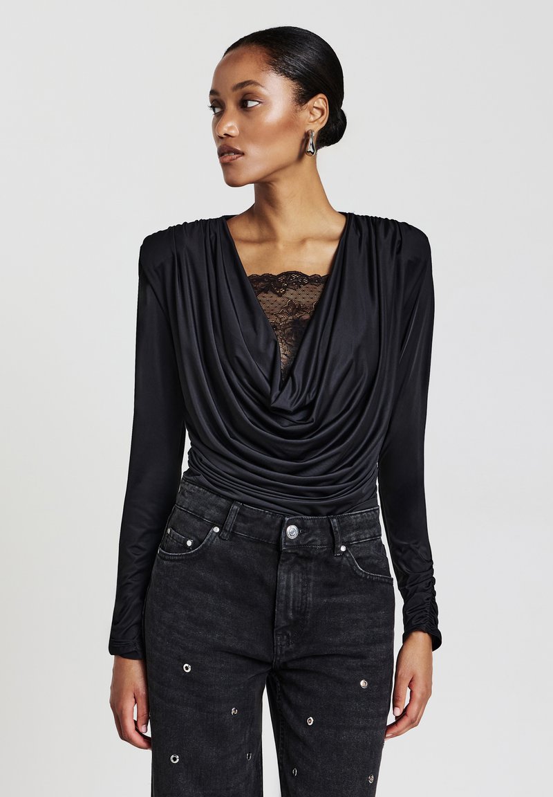 Schwarzes Langarmoberteil mit drapiertem Ausschnitt und transparentem Spitzen-Detail, kombiniert mit hoch taillierten schwarzen Jeans mit metallischen Akzenten.