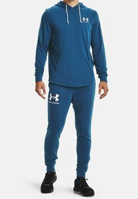 Completo sportivo blu composto da felpa con cappuccio e pantaloni realizzato in tessuto morbido. Presenta cappuccio, tasca frontale e accenti bianchi del logo. Indossato con scarpe da ginnastica nere.