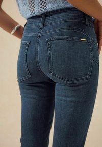 Los jeans de mezclilla azul oscuro presentan una cintura alta y dos bolsillos traseros. La tela tiene una textura suave con sutiles acentos de costura.