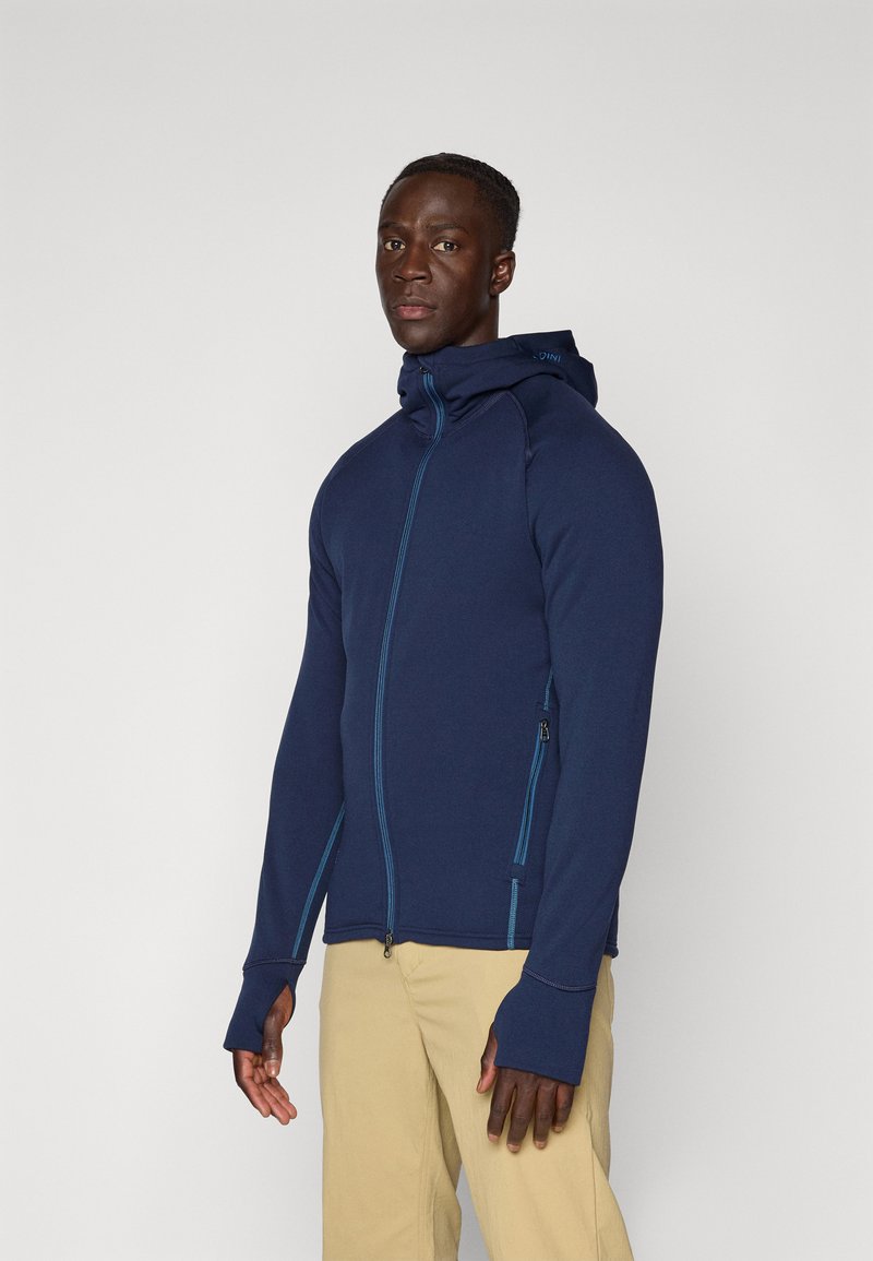 Navy zip-hoodie med kontrasterende turkisk syninger, hætte og fingerløse manchetter. Glat, strækbart stof; sidelomme med lynlås synlig.