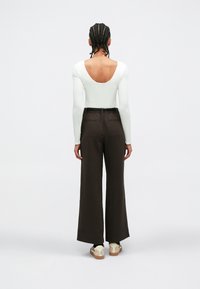 Long-sleeve white bodysuit with a scooped back, paired with high-waisted, wide-leg brown trousers. Braided hair and casual footwear visible.