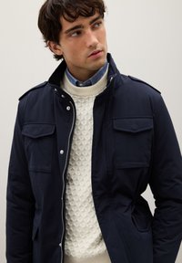 Giacca blu navy con due tasche sul petto e cerniera, indossata sopra un maglione crema a maglia. Presenta un colletto alto e una lucentezza sottile.
